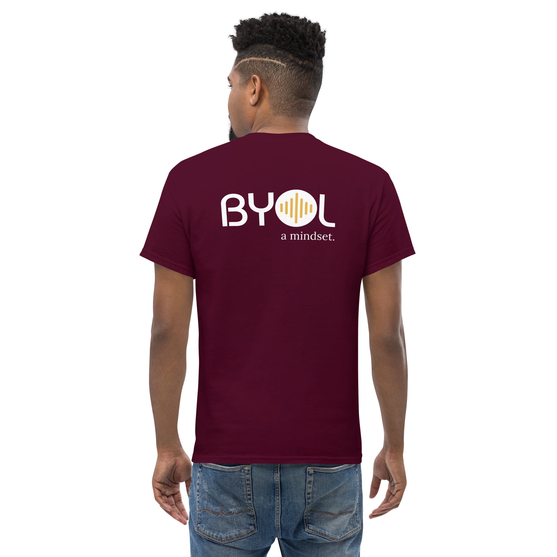 A young man with short curly hair wearing an maroon "BYOL: a mindset" T-shirt, viewed from the back. The T-shirt features the "BYOL" logo in white and yellow on the back and is available in multiple colors (maroon, black, brown, gold, plus more) and sizes (S-3XL). The "be you out loud" logo is displayed at the top.