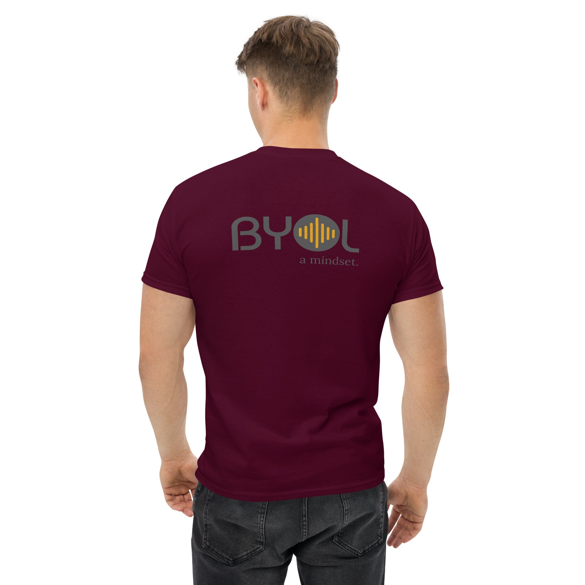 A young man with short blonde hair wearing a maroon "BYOL: a mindset" T-shirt, viewed from the back. The T-shirt features the "BYOL" logo in gray and yellow on the back and is available in multiple colors (maroon, black, brown, gold, plus more) and sizes (S-3XL). The "be you out loud" logo is displayed at the top.