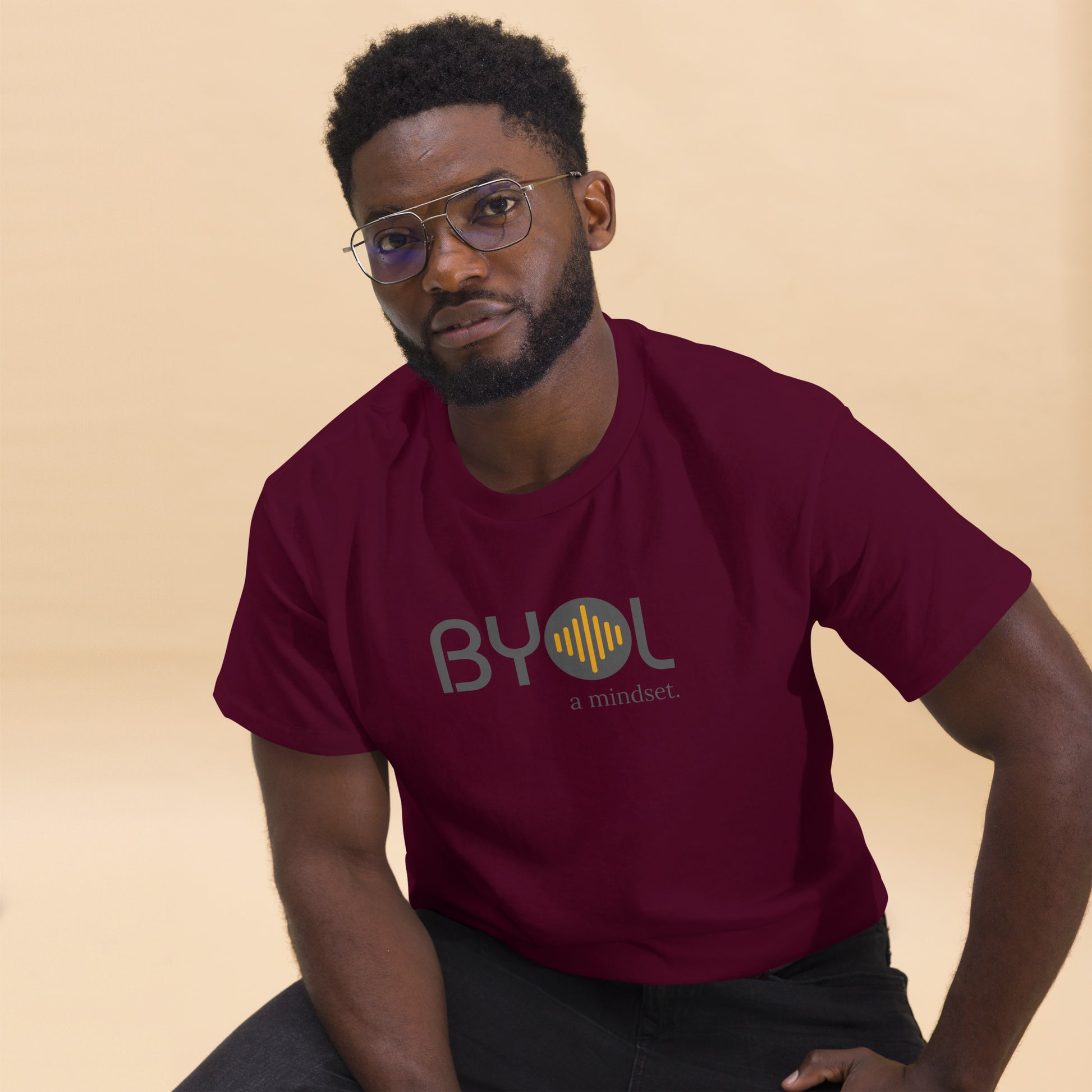 A young man with short curly hair and glasses wearing a maroon "BYOL: a mindset" T-shirt, sitting down. The T-shirt features the "BYOL" logo in gray and yellow on the front and is available in multiple colors (maroon, black, brown, gold, plus more) and sizes (S-3XL). The "be you out loud" logo is displayed at the top.