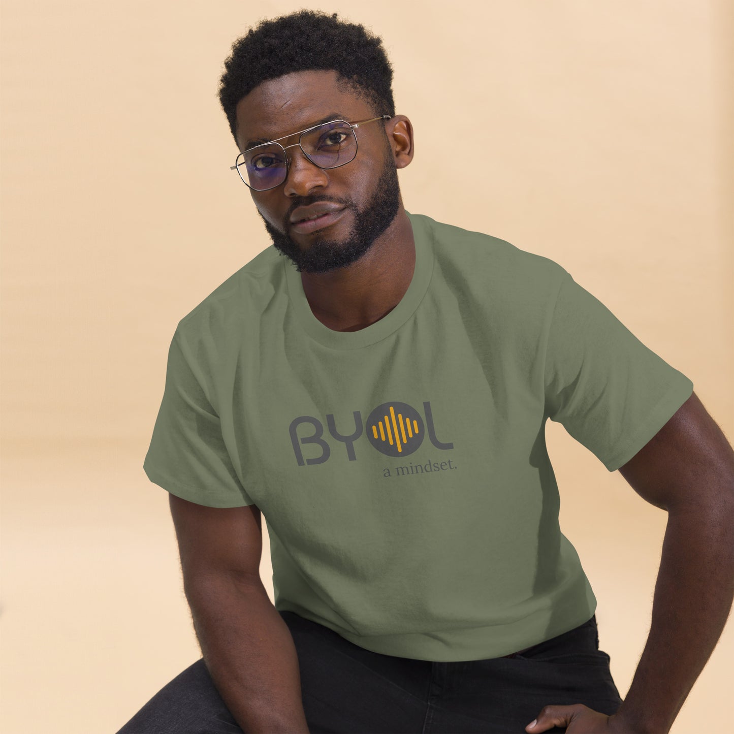 A young man with short curly hair and glasses wearing a military green "BYOL: a mindset" T-shirt, sitting down. The T-shirt features the "BYOL" logo in gray and yellow on the front and is available in multiple colors (maroon, black, brown, gold, plus more) and sizes (S-3XL). The "be you out loud" logo is displayed at the top.