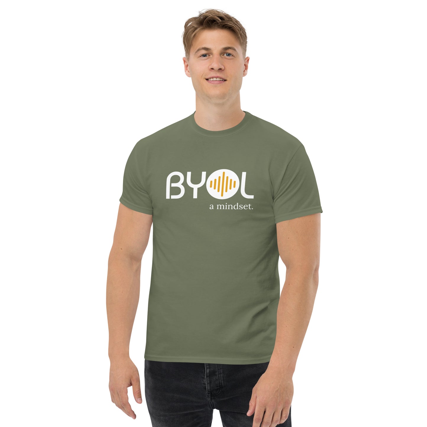 A young man with short blonde hair wearing an military green "BYOL: a mindset" T-shirt, smiling and standing with his arms at his sides. The T-shirt features the "BYOL" logo in white and yellow on the front and is available in multiple colors (maroon, black, brown, gold, plus more) and sizes (S-3XL). The "be you out loud" logo is displayed at the top.