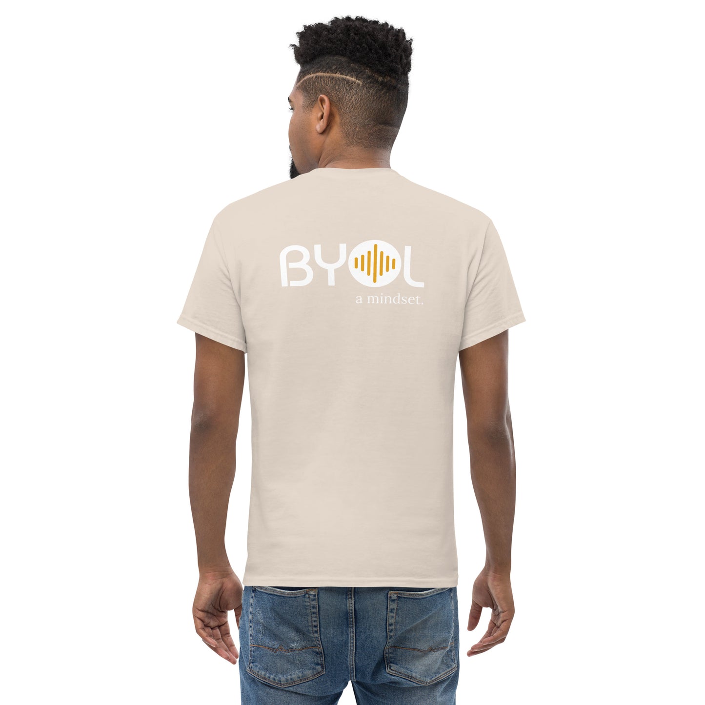 A young man with short curly hair wearing an sand "BYOL: a mindset" T-shirt, viewed from the back. The T-shirt features the "BYOL" logo in white and yellow on the back and is available in multiple colors (maroon, black, brown, gold, plus more) and sizes (S-3XL). The "be you out loud" logo is displayed at the top.