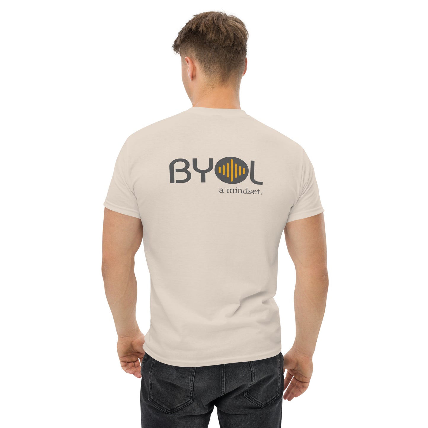 A young man with short blonde hair wearing a natural "BYOL: a mindset" T-shirt, viewed from the back. The T-shirt features the "BYOL" logo in gray and yellow on the back and is available in multiple colors (maroon, black, brown, gold, plus more) and sizes (S-3XL). The "be you out loud" logo is displayed at the top.