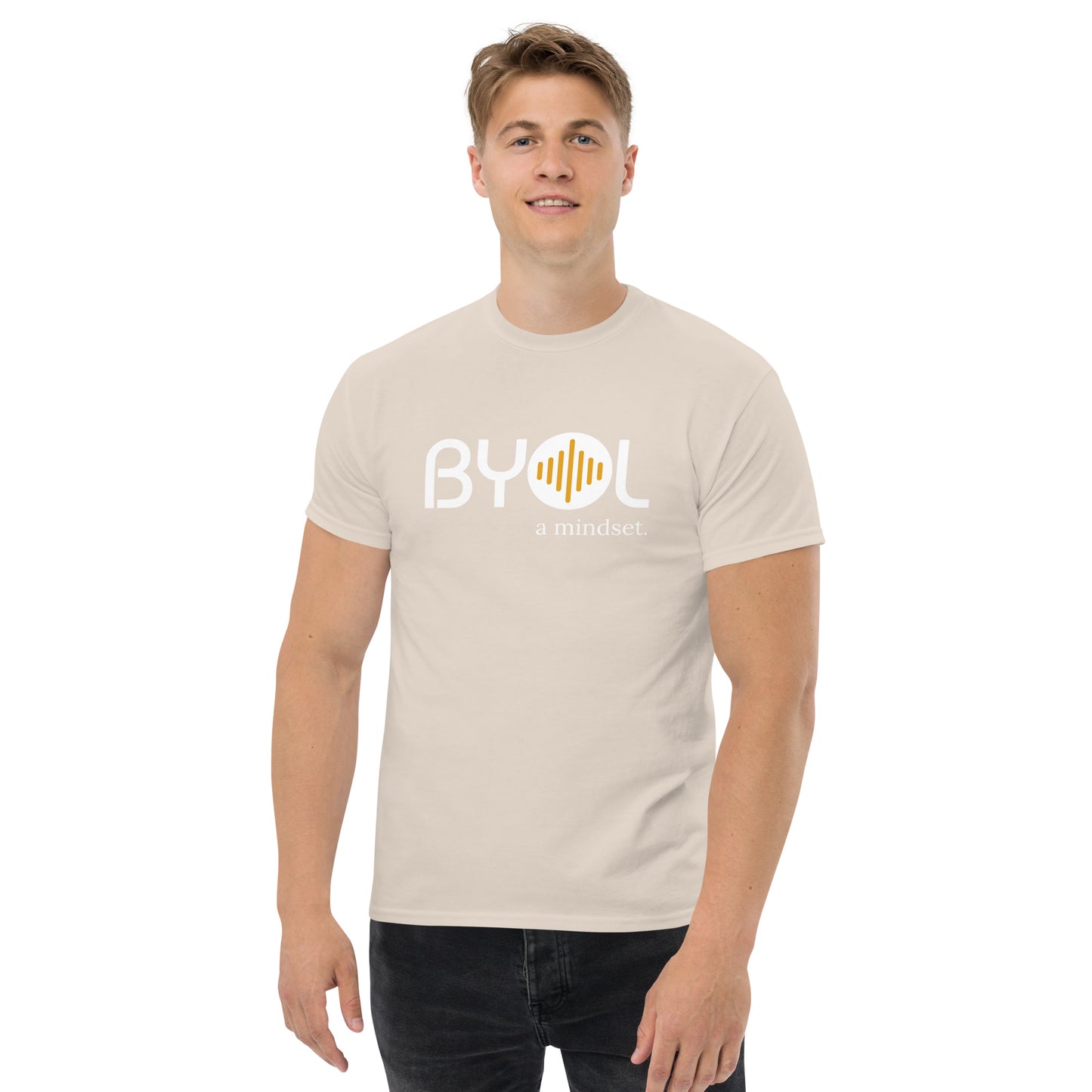 A young man with short blonde hair wearing an natural "BYOL: a mindset" T-shirt, smiling and standing with his arms at his sides. The T-shirt features the "BYOL" logo in white and yellow on the front and is available in multiple colors (maroon, black, brown, gold, plus more) and sizes (S-3XL). The "be you out loud" logo is displayed at the top.