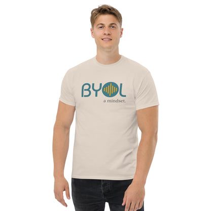 A young man with short blonde hair wearing a natural "BYOL: a mindset" T-shirt, smiling and standing with his arms at his sides. The T-shirt features the "BYOL" logo in teal and yellow on the front and is available in multiple colors (maroon, black, brown, gold, plus more) and sizes (S-3XL). The "be you out loud" logo is displayed at the top.