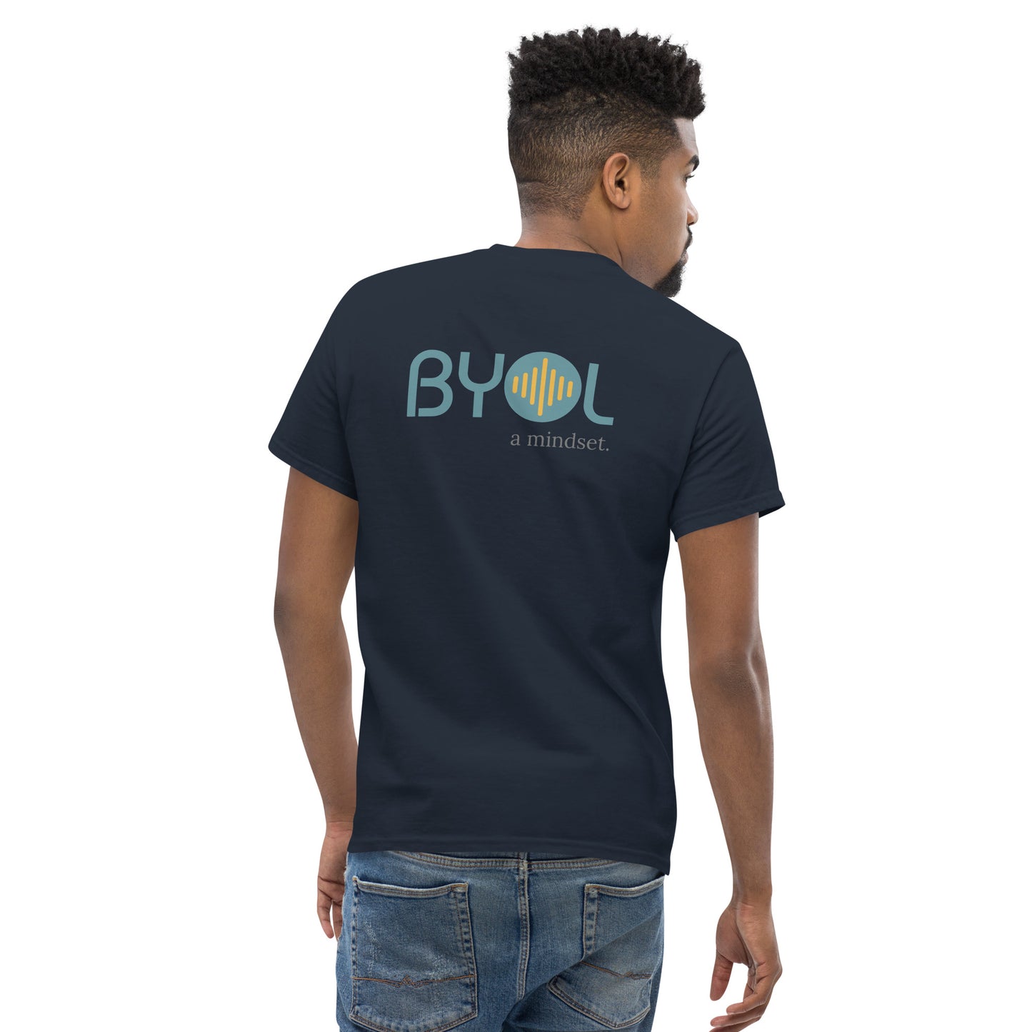 A young man with short hair wearing a navy blue "BYOL: a mindset" T-shirt, viewed from the back. The T-shirt features the "BYOL" logo in teal and yellow on the back and is available in multiple colors (maroon, black, brown, gold, plus more) and sizes (S-3XL). The "be you out loud" logo is displayed at the top.