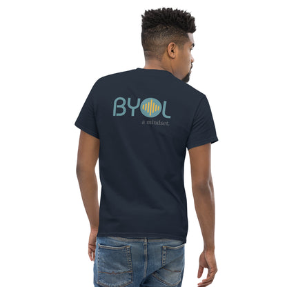 A young man with short hair wearing a navy blue "BYOL: a mindset" T-shirt, viewed from the back. The T-shirt features the "BYOL" logo in teal and yellow on the back and is available in multiple colors (maroon, black, brown, gold, plus more) and sizes (S-3XL). The "be you out loud" logo is displayed at the top.