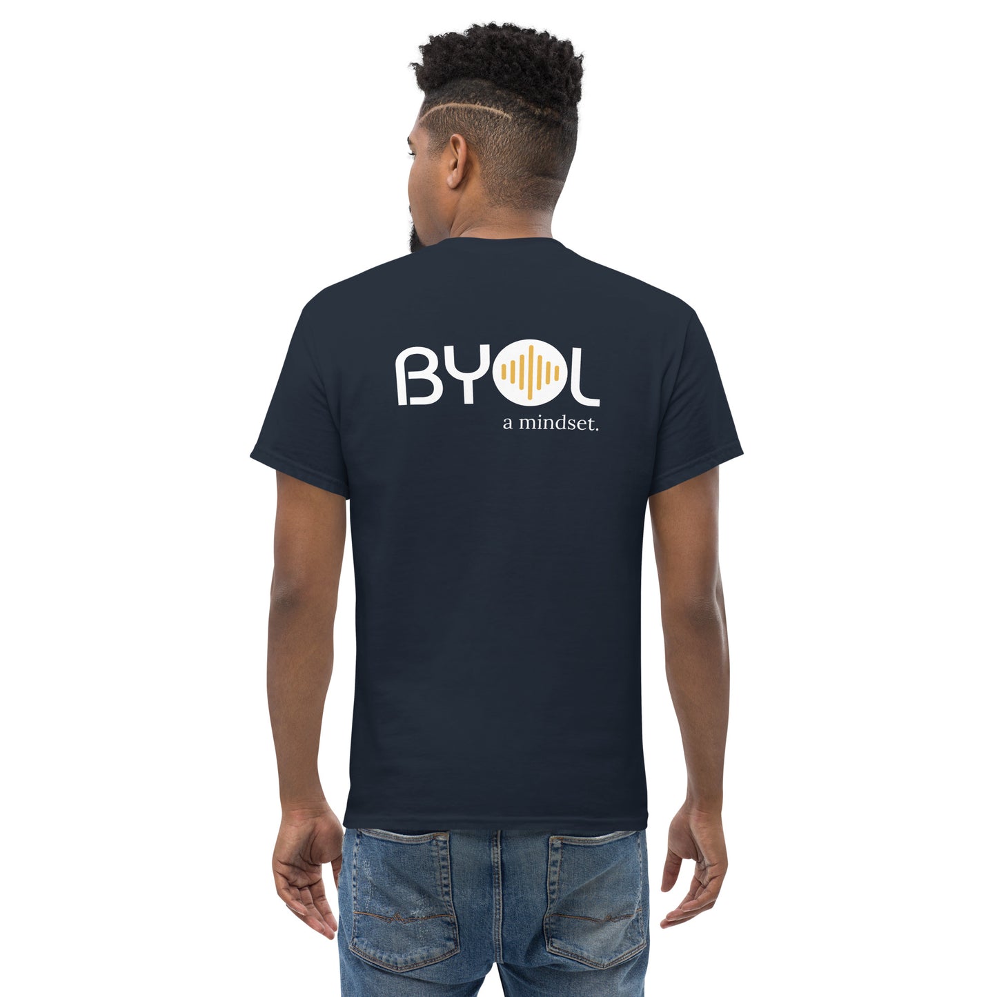 A young man with short curly hair wearing an navy blue "BYOL: a mindset" T-shirt, viewed from the back. The T-shirt features the "BYOL" logo in white and yellow on the back and is available in multiple colors (maroon, black, brown, gold, plus more) and sizes (S-3XL). The "be you out loud" logo is displayed at the top.