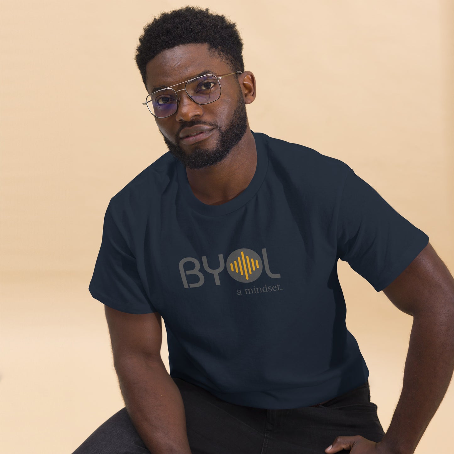 A young man with short curly hair and glasses wearing a navy blue "BYOL: a mindset" T-shirt, sitting down. The T-shirt features the "BYOL" logo in gray and yellow on the front and is available in multiple colors (maroon, black, brown, gold, plus more) and sizes (S-3XL). The "be you out loud" logo is displayed at the top.