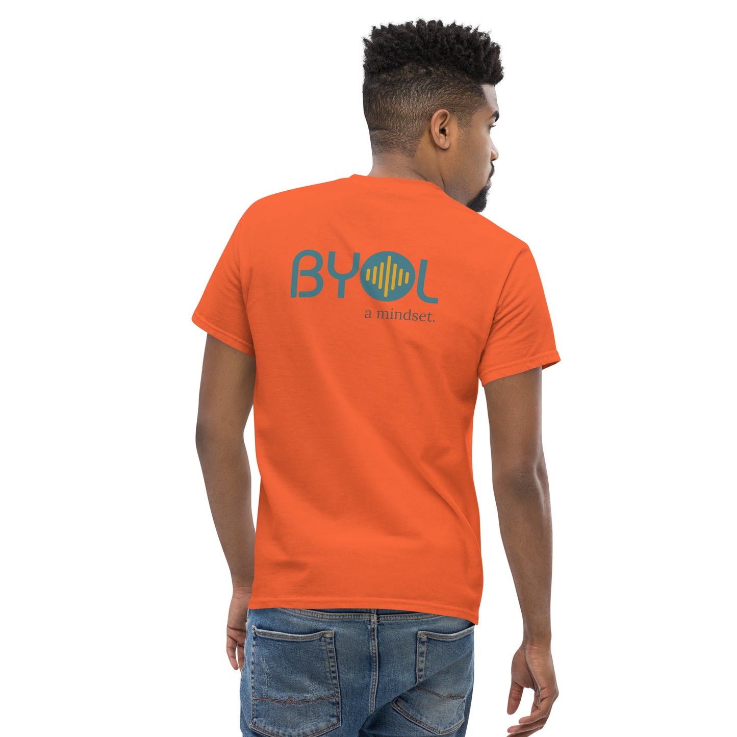 A young man with short hair wearing a orange "BYOL: a mindset" T-shirt, viewed from the back. The T-shirt features the "BYOL" logo in teal and yellow on the back and is available in multiple colors (maroon, black, brown, gold, plus more) and sizes (S-3XL). The "be you out loud" logo is displayed at the top.