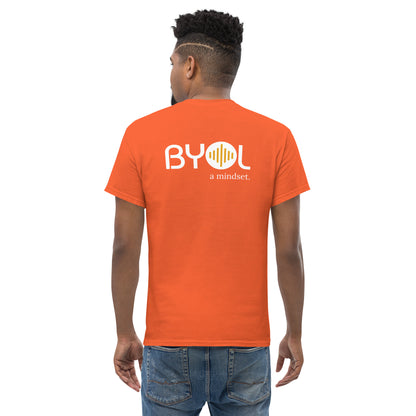 A young man with short curly hair wearing an orange "BYOL: a mindset" T-shirt, viewed from the back. The T-shirt features the "BYOL" logo in white and yellow on the back and is available in multiple colors (maroon, black, brown, gold, plus more) and sizes (S-3XL). The "be you out loud" logo is displayed at the top.