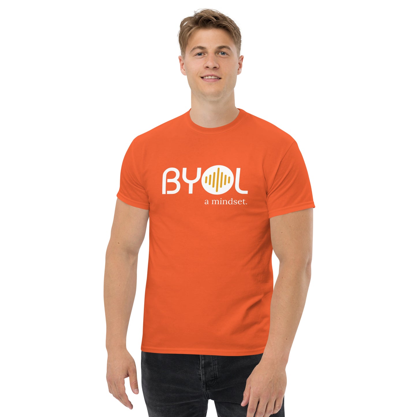 A young man with short blonde hair wearing an orange "BYOL: a mindset" T-shirt, smiling and standing with his arms at his sides. The T-shirt features the "BYOL" logo in white and yellow on the front and is available in multiple colors (maroon, black, brown, gold, plus more) and sizes (S-3XL). The "be you out loud" logo is displayed at the top.