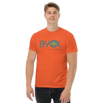 A young man with short blonde hair wearing a orange "BYOL: a mindset" T-shirt, smiling and standing with his arms at his sides. The T-shirt features the "BYOL" logo in teal and yellow on the front and is available in multiple colors (maroon, black, brown, gold, plus more) and sizes (S-3XL). The "be you out loud" logo is displayed at the top.