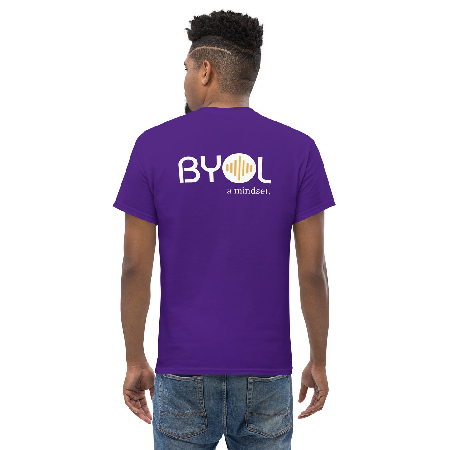 A young man with short curly hair wearing an purple "BYOL: a mindset" T-shirt, viewed from the back. The T-shirt features the "BYOL" logo in white and yellow on the back and is available in multiple colors (maroon, black, brown, gold, plus more) and sizes (S-3XL). The "be you out loud" logo is displayed at the top.