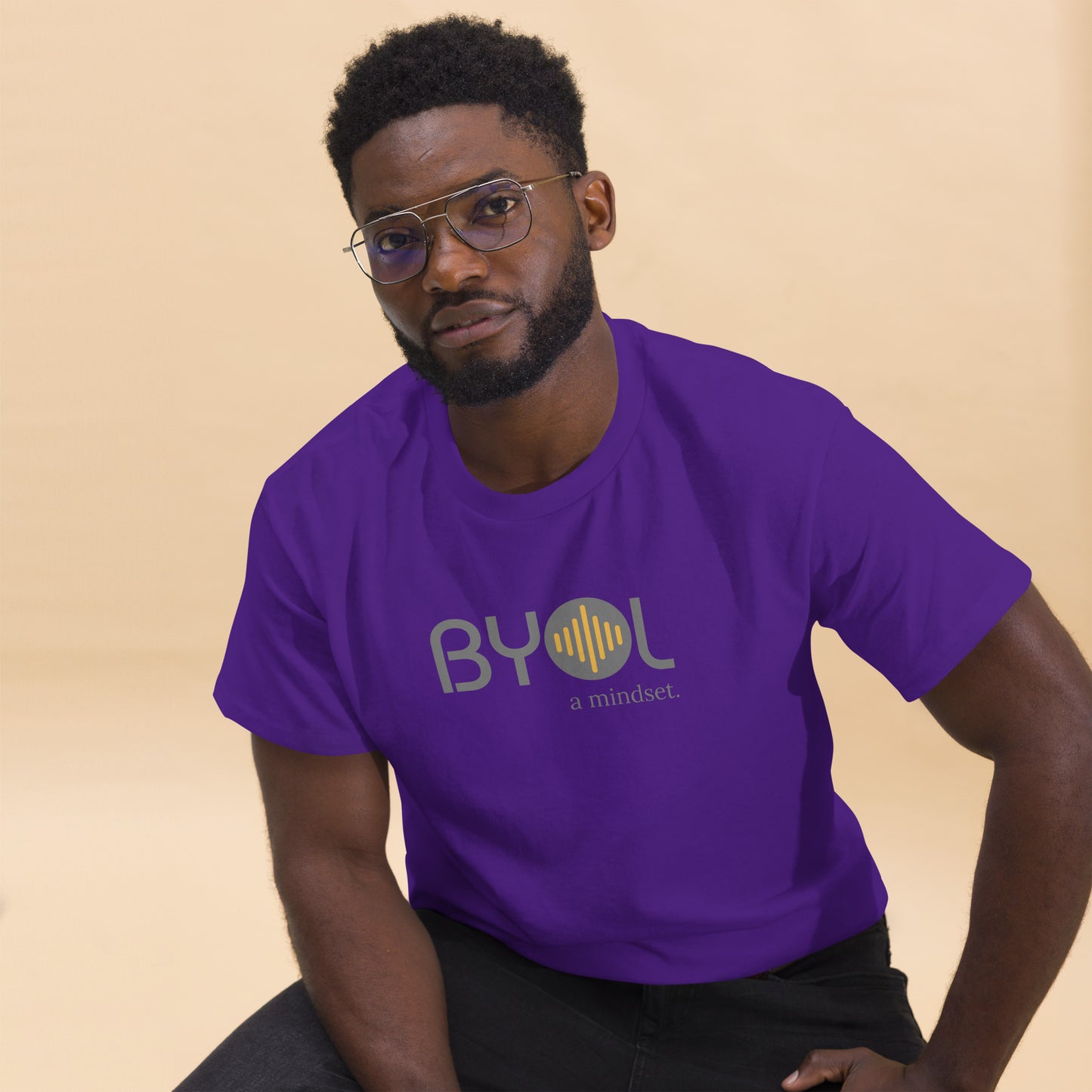 A young man with short curly hair and glasses wearing a purple "BYOL: a mindset" T-shirt, sitting down. The T-shirt features the "BYOL" logo in gray and yellow on the front and is available in multiple colors (maroon, black, brown, gold, plus more) and sizes (S-3XL). The "be you out loud" logo is displayed at the top.