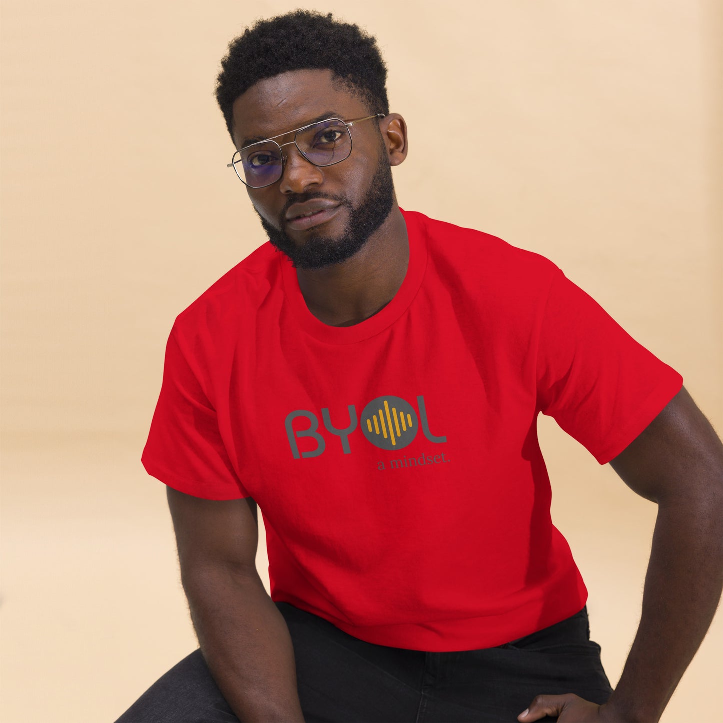 A young man with short curly hair and glasses wearing a red "BYOL: a mindset" T-shirt, sitting down. The T-shirt features the "BYOL" logo in gray and yellow on the front and is available in multiple colors (maroon, black, brown, gold, plus more) and sizes (S-3XL). The "be you out loud" logo is displayed at the top.