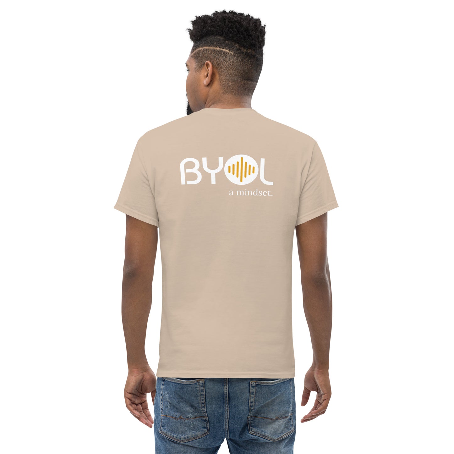 A young man with short curly hair wearing an natural "BYOL: a mindset" T-shirt, viewed from the back. The T-shirt features the "BYOL" logo in white and yellow on the back and is available in multiple colors (maroon, black, brown, gold, plus more) and sizes (S-3XL). The "be you out loud" logo is displayed at the top.