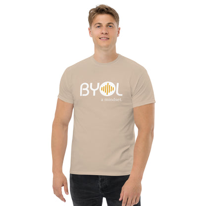 A young man with short blonde hair wearing an sand "BYOL: a mindset" T-shirt, smiling and standing with his arms at his sides. The T-shirt features the "BYOL" logo in white and yellow on the front and is available in multiple colors (maroon, black, brown, gold, plus more) and sizes (S-3XL). The "be you out loud" logo is displayed at the top.