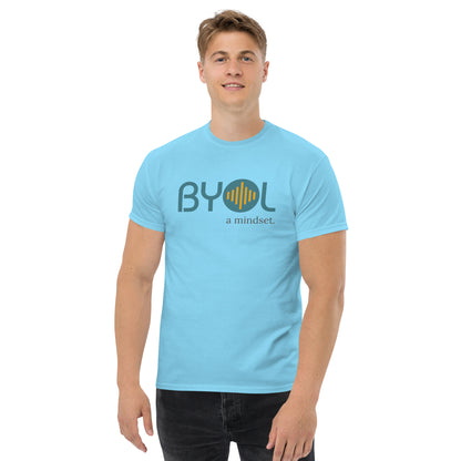 A young man with short blonde hair wearing a sky "BYOL: a mindset" T-shirt, smiling and standing with his arms at his sides. The T-shirt features the "BYOL" logo in teal and yellow on the front and is available in multiple colors (maroon, black, brown, gold, plus more) and sizes (S-3XL). The "be you out loud" logo is displayed at the top.