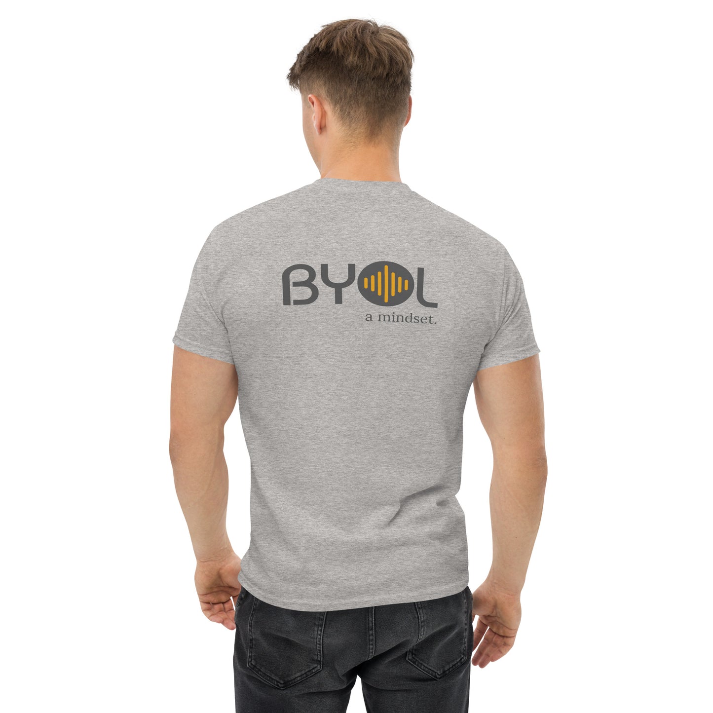 A young man with short blonde hair wearing a ash "BYOL: a mindset" T-shirt, viewed from the back. The T-shirt features the "BYOL" logo in gray and yellow on the back and is available in multiple colors (maroon, black, brown, gold, plus more) and sizes (S-3XL). The "be you out loud" logo is displayed at the top.