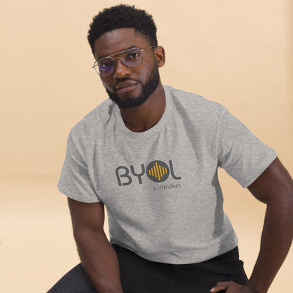 A young man with short curly hair and glasses wearing a ash "BYOL: a mindset" T-shirt, sitting down. The T-shirt features the "BYOL" logo in gray and yellow on the front and is available in multiple colors (maroon, black, brown, gold, plus more) and sizes (S-3XL). The "be you out loud" logo is displayed at the top.