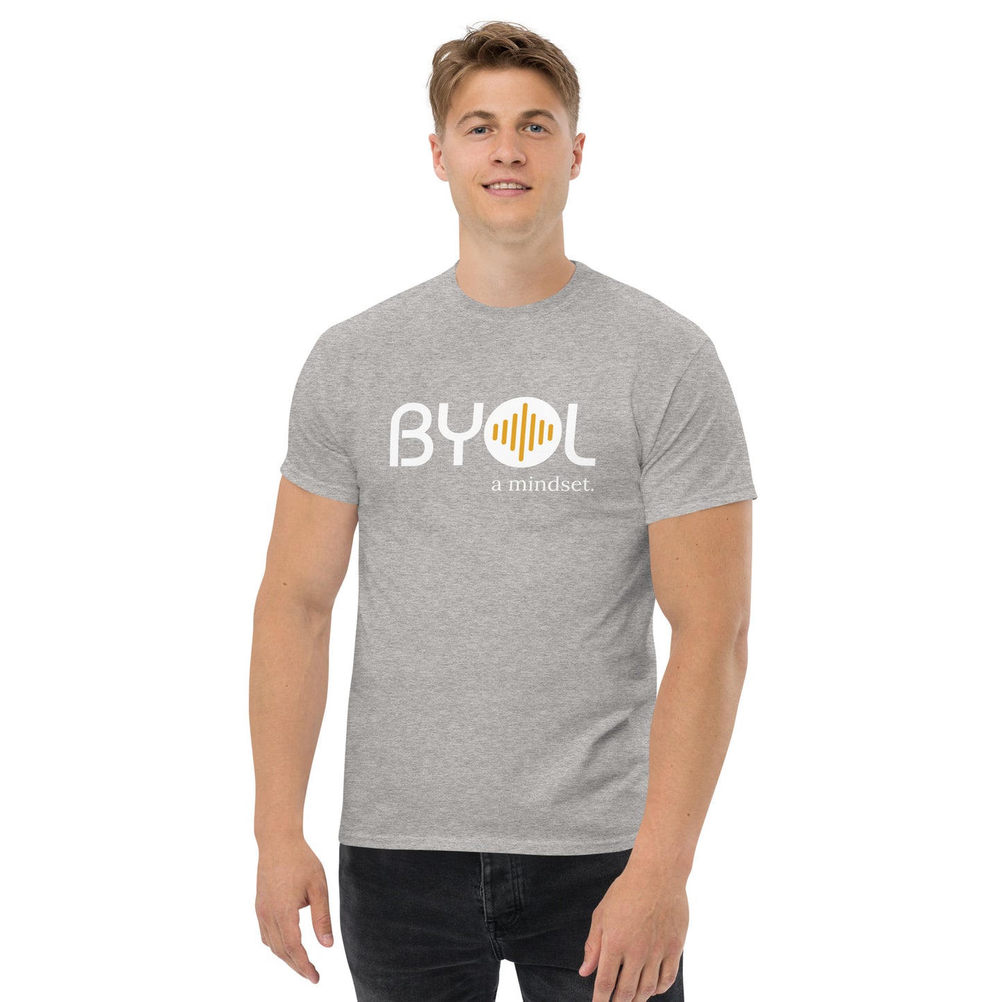 A young man with short blonde hair wearing an ash "BYOL: a mindset" T-shirt, smiling and standing with his arms at his sides. The T-shirt features the "BYOL" logo in white and yellow on the front and is available in multiple colors (maroon, black, brown, gold, plus more) and sizes (S-3XL). The "be you out loud" logo is displayed at the top.