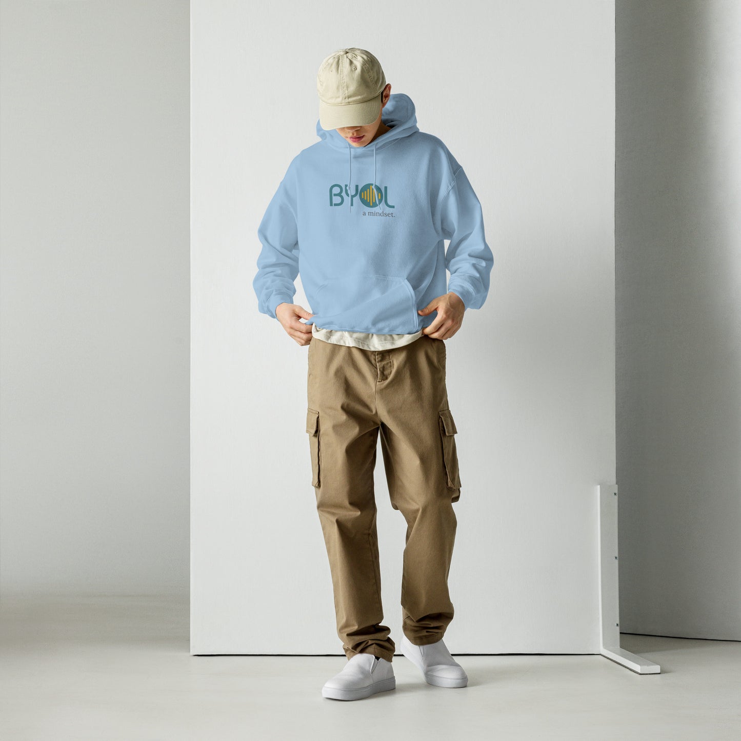 A young man wearing a light blue "BYOL: a mindset" hoodie, adjusting the hem and looking down. He is also wearing a beige cap and khaki pants. The hoodie features the "BYOL" logo in teal and yellow on the front and is available in multiple colors (black, maroon, green, beige, plus more) and sizes (S-3XL). The "be you out loud" logo is displayed at the top.