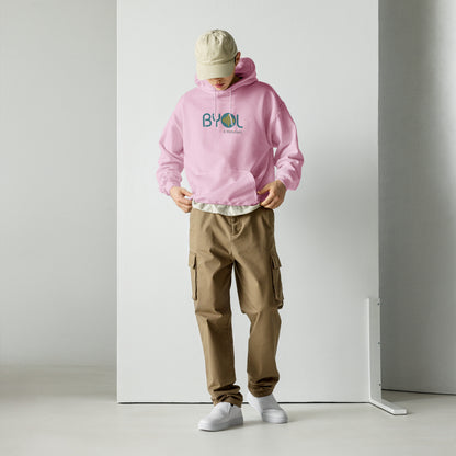 A young man wearing a pink "BYOL: a mindset" hoodie, adjusting the hem and looking down. He is also wearing a beige cap and khaki pants. The hoodie features the "BYOL" logo in teal and yellow on the front and is available in multiple colors (black, maroon, green, beige, plus more) and sizes (S-3XL). The "be you out loud" logo is displayed at the top.