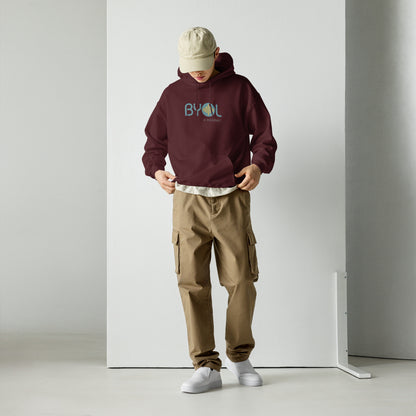 A young man wearing a maroon "BYOL: a mindset" hoodie, adjusting the hem and looking down. He is also wearing a beige cap and khaki pants. The hoodie features the "BYOL" logo in teal and yellow on the front and is available in multiple colors (black, maroon, green, beige, plus more) and sizes (S-3XL). The "be you out loud" logo is displayed at the top.