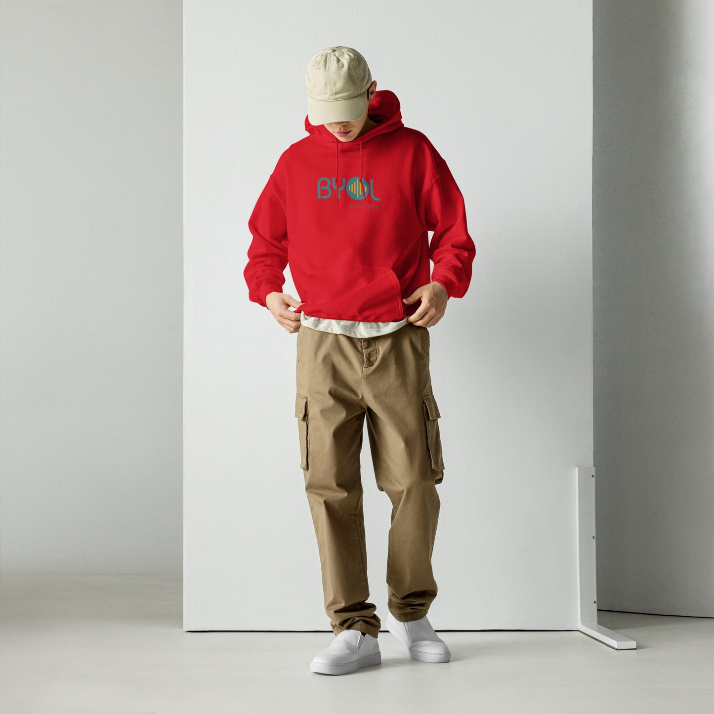 A young man wearing a red "BYOL: a mindset" hoodie, adjusting the hem and looking down. He is also wearing a beige cap and khaki pants. The hoodie features the "BYOL" logo in teal and yellow on the front and is available in multiple colors (black, maroon, green, beige, plus more) and sizes (S-3XL). The "be you out loud" logo is displayed at the top.