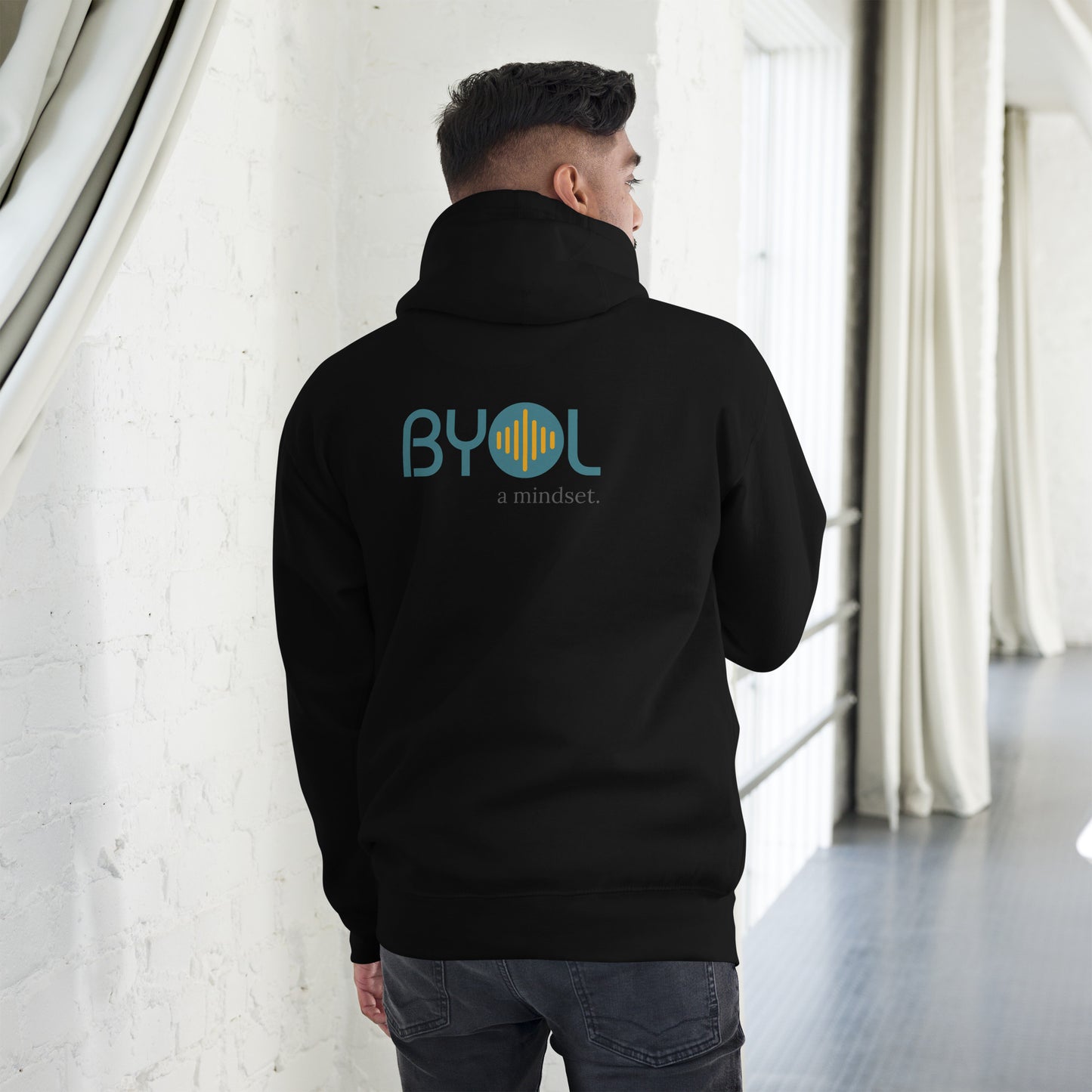 A young man wearing a black "BYOL: a mindset" hoodie, viewed from the back. The hoodie features the "BYOL" logo in teal and yellow on the back and is available in multiple colors (black, red, maroon, brown, plus more) and sizes (S-3XL). The "be you out loud" logo is displayed at the top.