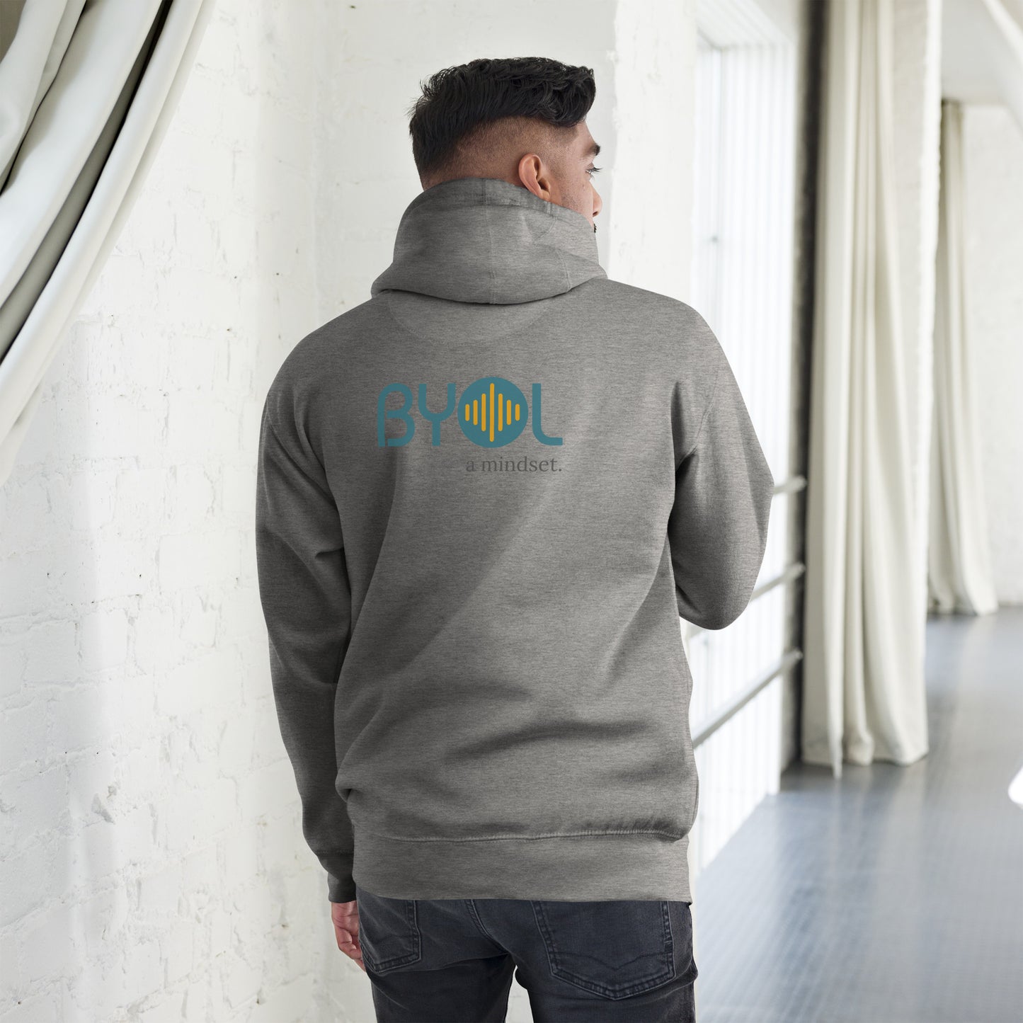 A young man wearing a heather grey "BYOL: a mindset" hoodie, viewed from the back. The hoodie features the "BYOL" logo in teal and yellow on the back and is available in multiple colors (black, red, maroon, brown, plus more) and sizes (S-3XL). The "be you out loud" logo is displayed at the top.