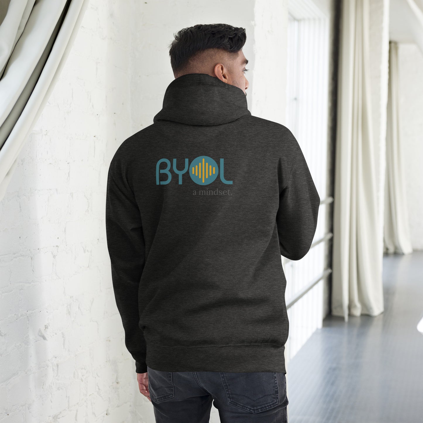 A young man wearing a dark gray "BYOL: a mindset" hoodie, viewed from the back. The hoodie features the "BYOL" logo in teal and yellow on the back and is available in multiple colors (black, red, maroon, brown, plus more) and sizes (S-3XL). The "be you out loud" logo is displayed at the top.