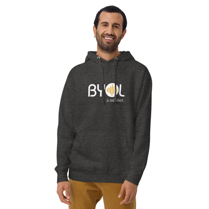 A young man with short hair and a beard wearing a dark heather grey "BYOL: a mindset" hoodie, smiling and standing with his arms at his sides. The hoodie features the "BYOL" logo in white and yellow on the front and is available in multiple colors (black, dark gray, maroon, pink, light gray, plus more) and sizes (S-3XL). The "be you out loud" logo is displayed at the top.