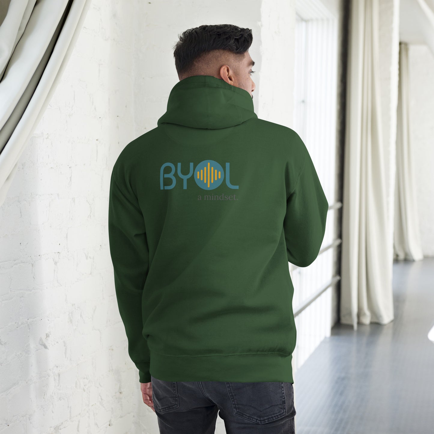 A young man wearing a green "BYOL: a mindset" hoodie, viewed from the back. The hoodie features the "BYOL" logo in teal and yellow on the back and is available in multiple colors (black, red, maroon, brown, plus more) and sizes (S-3XL). The "be you out loud" logo is displayed at the top.