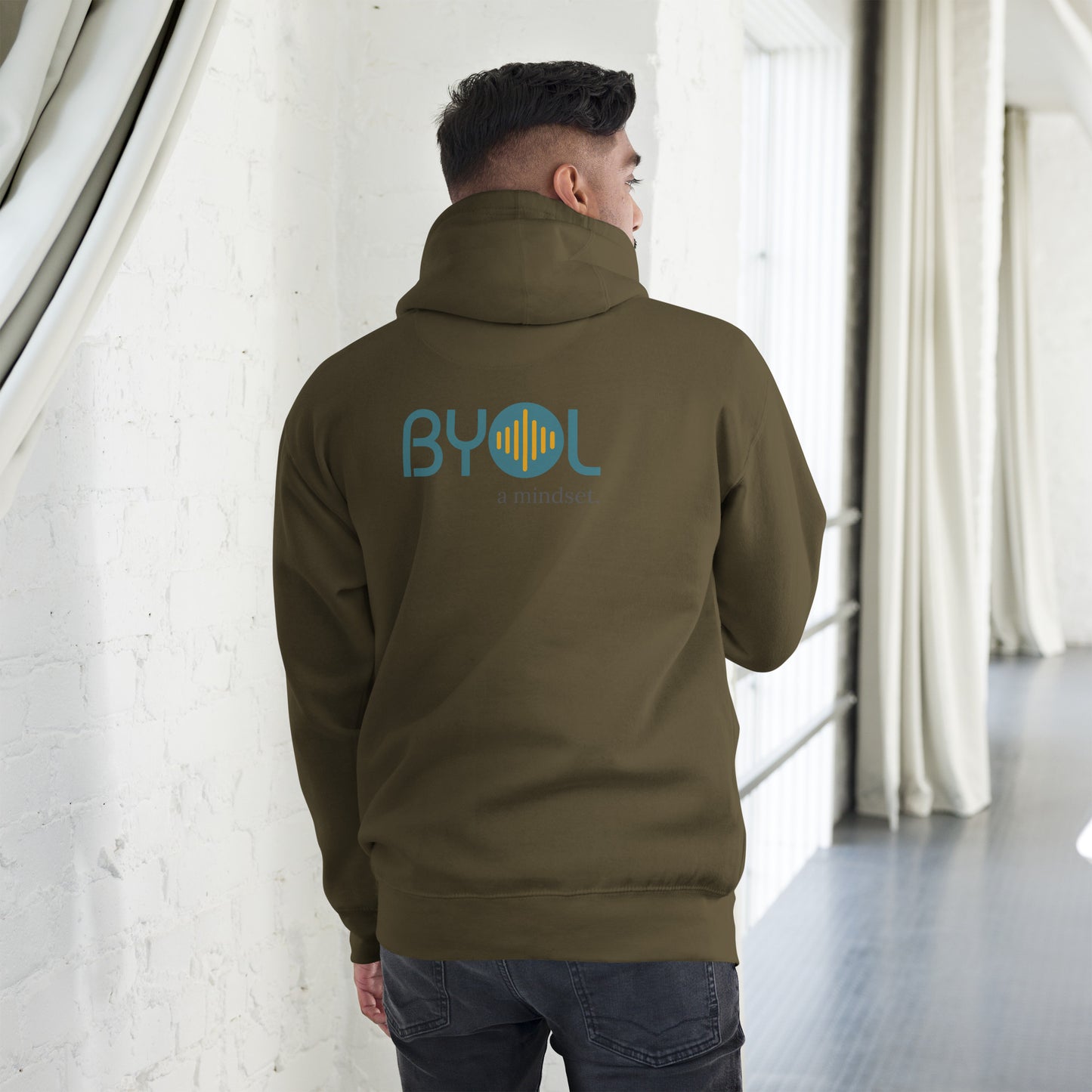 A young man wearing a military green "BYOL: a mindset" hoodie, viewed from the back. The hoodie features the "BYOL" logo in teal and yellow on the back and is available in multiple colors (black, red, maroon, brown, plus more) and sizes (S-3XL). The "be you out loud" logo is displayed at the top.