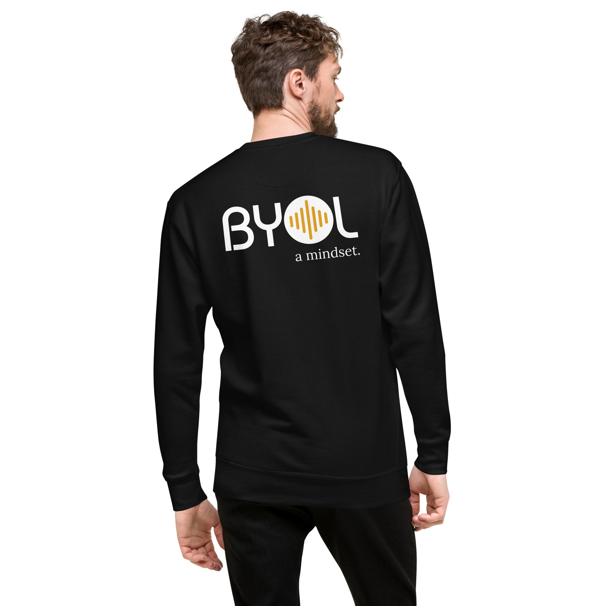 A young man with short hair and a beard wearing a black "BYOL: a mindset" sweatshirt, viewed from the back. The sweatshirt features the "BYOL" logo in white and yellow on the back and is available in multiple colors (black, dark gray, light gray, pink, plus more) and sizes (S-3XL). The "be you out loud" logo is displayed at the top.