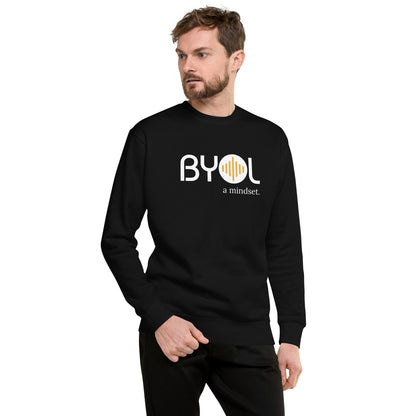 A young man with short hair and a beard wearing a black "BYOL: a mindset" sweatshirt, standing and looking to the side. The sweatshirt features the "BYOL" logo in white and yellow on the front and is available in multiple colors (black, dark gray, light gray, pink, plus more) and sizes (S-3XL). The "be you out loud" logo is displayed at the top.