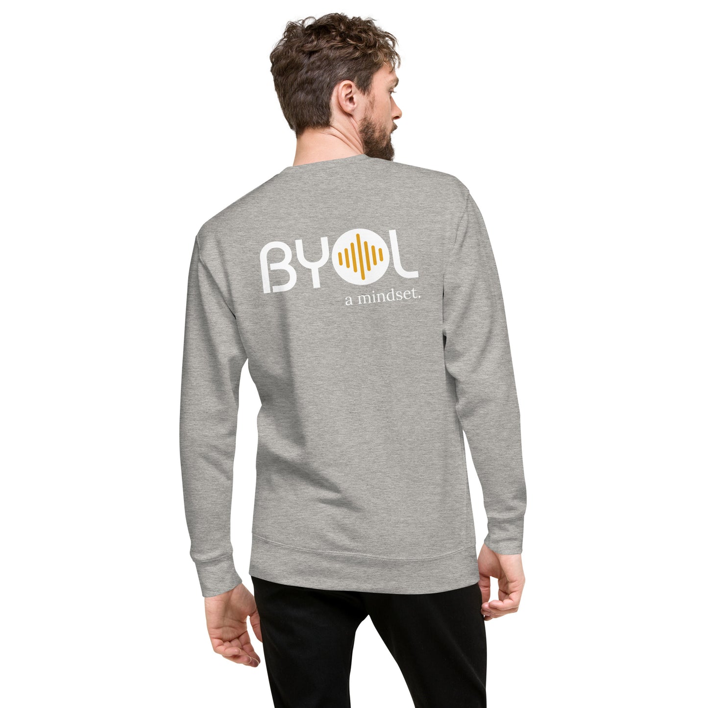 A young man with short hair and a beard wearing a light heather grey "BYOL: a mindset" sweatshirt, viewed from the back. The sweatshirt features the "BYOL" logo in white and yellow on the back and is available in multiple colors (black, dark gray, light gray, pink, plus more) and sizes (S-3XL). The "be you out loud" logo is displayed at the top.