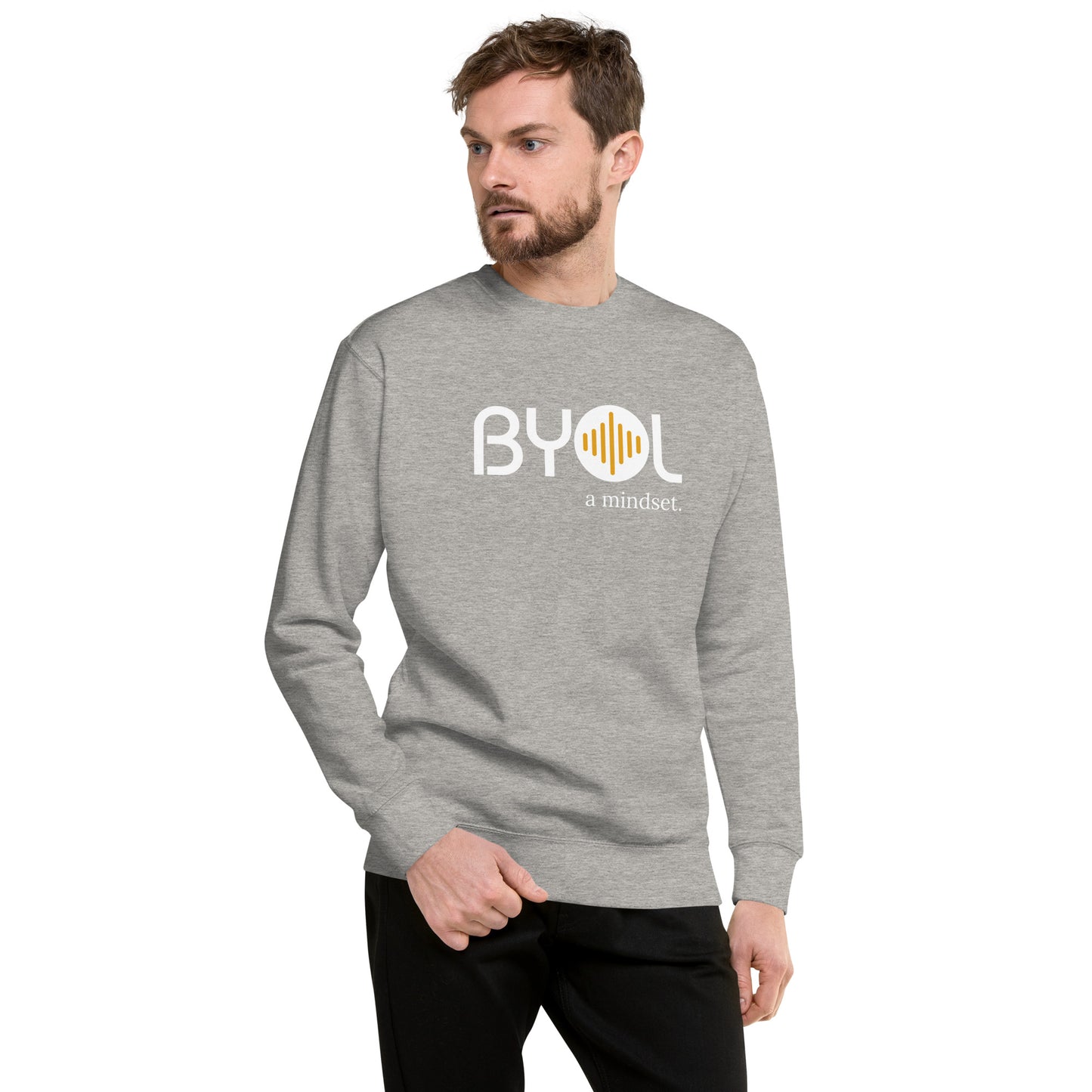 A young man with short hair and a beard wearing a carbon grey "BYOL: a mindset" sweatshirt, standing and looking to the side. The sweatshirt features the "BYOL" logo in white and yellow on the front and is available in multiple colors (black, dark gray, light gray, pink, plus more) and sizes (S-3XL). The "be you out loud" logo is displayed at the top.