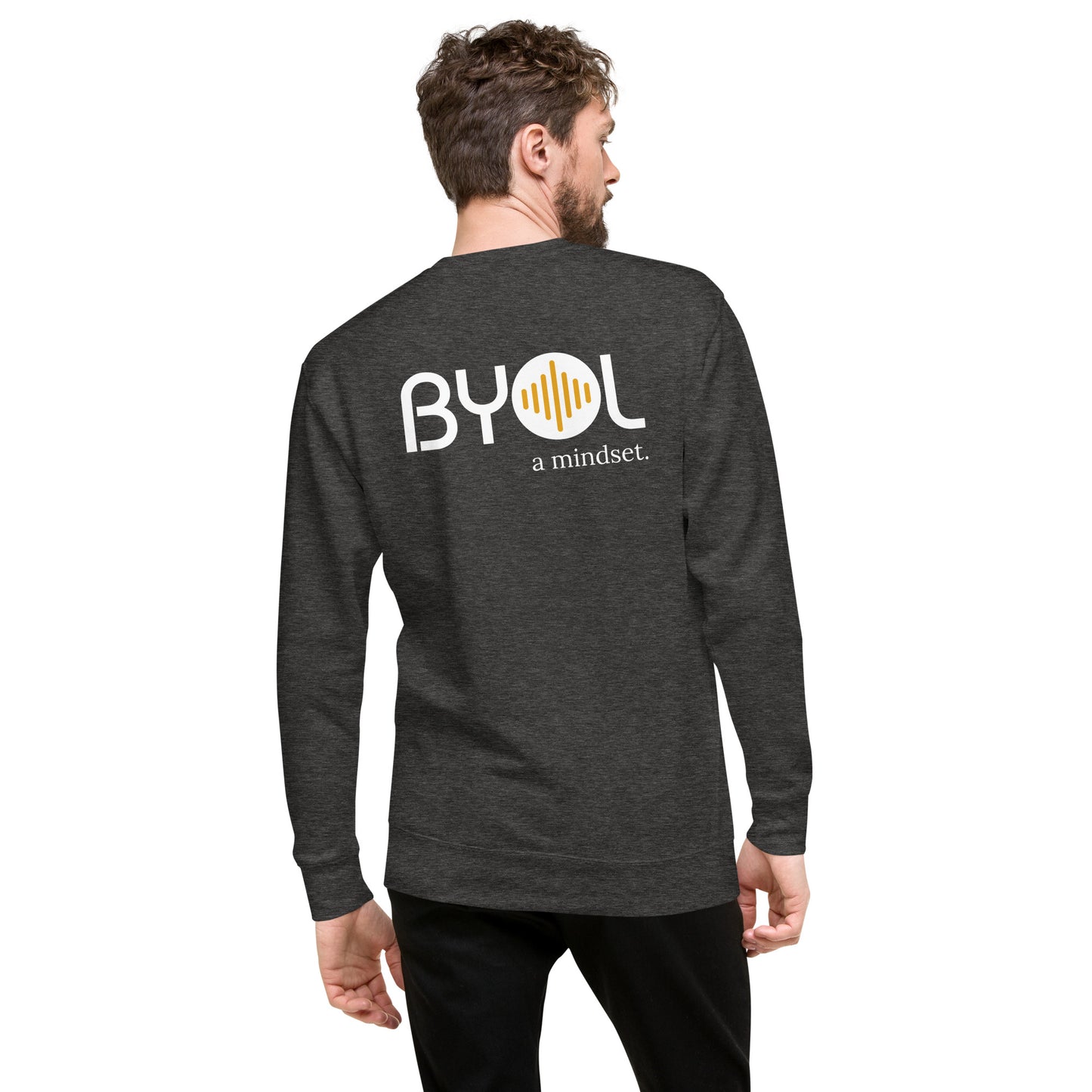 A young man with short hair and a beard wearing a dark heather grey "BYOL: a mindset" sweatshirt, viewed from the back. The sweatshirt features the "BYOL" logo in white and yellow on the back and is available in multiple colors (black, dark gray, light gray, pink, plus more) and sizes (S-3XL). The "be you out loud" logo is displayed at the top.