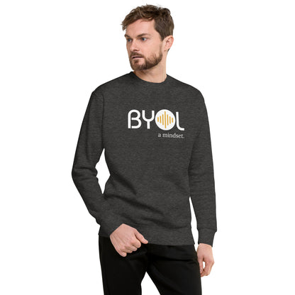 A young man with short hair and a beard wearing a dark heather grey "BYOL: a mindset" sweatshirt, standing and looking to the side. The sweatshirt features the "BYOL" logo in white and yellow on the front and is available in multiple colors (black, dark gray, light gray, pink, plus more) and sizes (S-3XL). The "be you out loud" logo is displayed at the top.