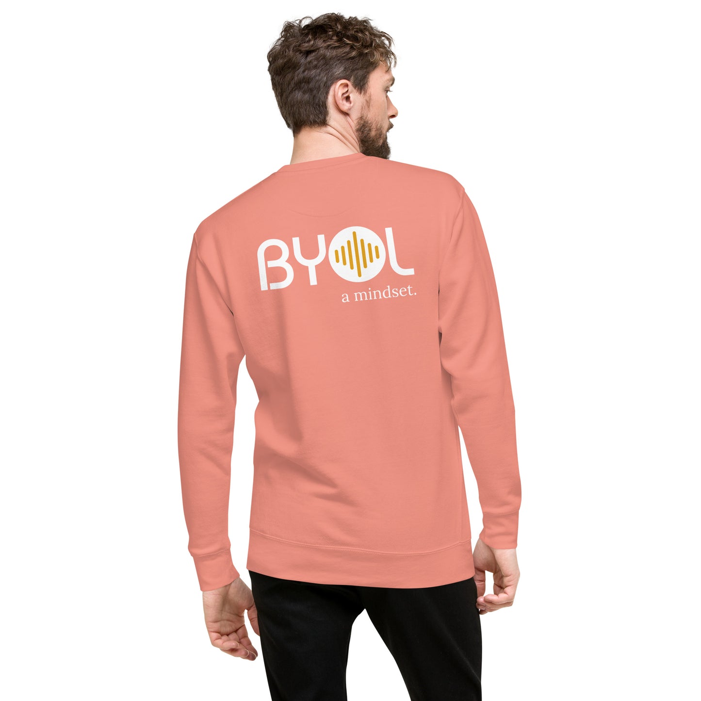 A young man with short hair and a beard wearing a dusty pink "BYOL: a mindset" sweatshirt, viewed from the back. The sweatshirt features the "BYOL" logo in white and yellow on the back and is available in multiple colors (black, dark gray, light gray, pink, plus more) and sizes (S-3XL). The "be you out loud" logo is displayed at the top.