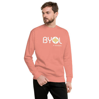 A young man with short hair and a beard wearing a dusty rose "BYOL: a mindset" sweatshirt, standing and looking to the side. The sweatshirt features the "BYOL" logo in white and yellow on the front and is available in multiple colors (black, dark gray, light gray, pink, plus more) and sizes (S-3XL). The "be you out loud" logo is displayed at the top.