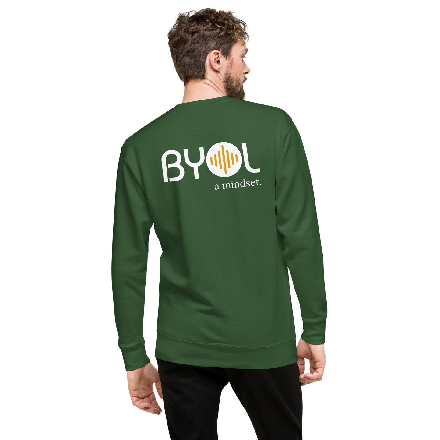 A young man with short hair and a beard wearing a green "BYOL: a mindset" sweatshirt, viewed from the back. The sweatshirt features the "BYOL" logo in white and yellow on the back and is available in multiple colors (black, dark gray, light gray, pink, plus more) and sizes (S-3XL). The "be you out loud" logo is displayed at the top.