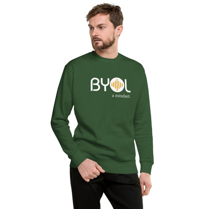 A young man with short hair and a beard wearing a green "BYOL: a mindset" sweatshirt, standing and looking to the side. The sweatshirt features the "BYOL" logo in white and yellow on the front and is available in multiple colors (black, dark gray, light gray, pink, plus more) and sizes (S-3XL). The "be you out loud" logo is displayed at the top.