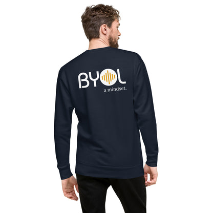 A young man with short hair and a beard wearing a navy blue "BYOL: a mindset" sweatshirt, viewed from the back. The sweatshirt features the "BYOL" logo in white and yellow on the back and is available in multiple colors (black, dark gray, light gray, pink, plus more) and sizes (S-3XL). The "be you out loud" logo is displayed at the top.