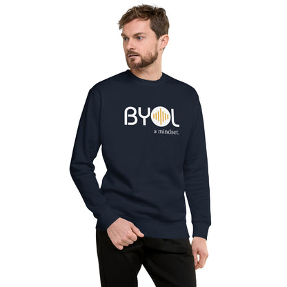 A young man with short hair and a beard wearing a navy blue "BYOL: a mindset" sweatshirt, standing and looking to the side. The sweatshirt features the "BYOL" logo in white and yellow on the front and is available in multiple colors (black, dark gray, light gray, pink, plus more) and sizes (S-3XL). The "be you out loud" logo is displayed at the top.