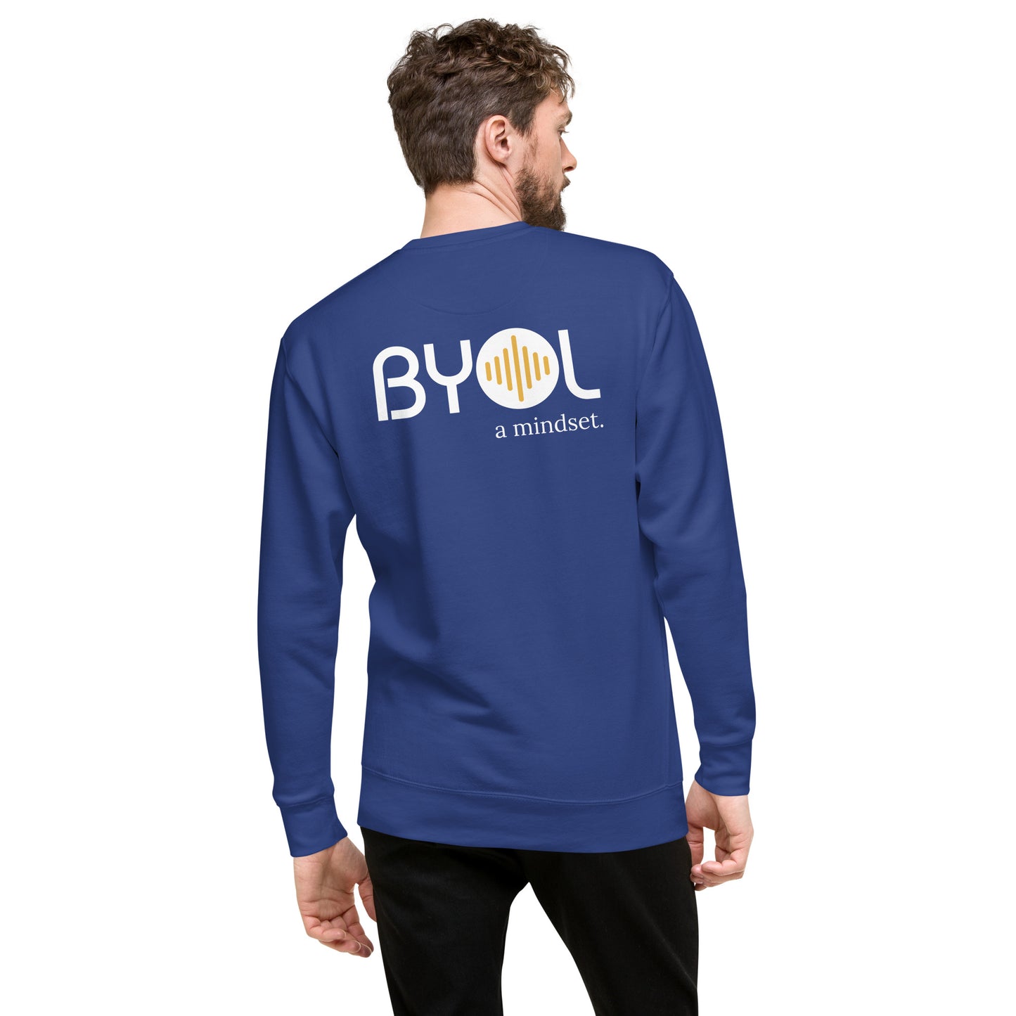 A young man with short hair and a beard wearing a royal blue "BYOL: a mindset" sweatshirt, viewed from the back. The sweatshirt features the "BYOL" logo in white and yellow on the back and is available in multiple colors (black, dark gray, light gray, pink, plus more) and sizes (S-3XL). The "be you out loud" logo is displayed at the top.