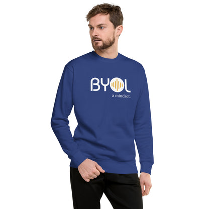 A young man with short hair and a beard wearing a royal blue "BYOL: a mindset" sweatshirt, standing and looking to the side. The sweatshirt features the "BYOL" logo in white and yellow on the front and is available in multiple colors (black, dark gray, light gray, pink, plus more) and sizes (S-3XL). The "be you out loud" logo is displayed at the top.